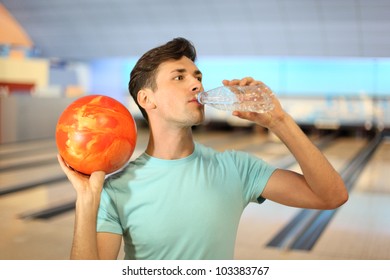 Young Man Holds Orange Ball And Drinks Pure Water From Bottle In Bowling Club; Shallow Depth Of Field