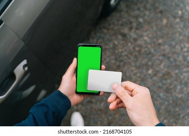 A Young Man Holds A Smartphone And A Bank Card In His Hands. Smartphone With A Green Screen. The Guy Is Standing Next To The Car.