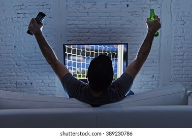 Young Man Home Alone Watching Soccer Or Football Game In Television Enjoying And Celebrating Goal And Victory Holding Beer Bottle And TV Controller Gesturing On The Sofa Happy And Excited