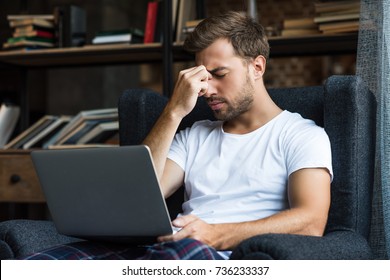 Young man in home clothes sitting in armchair with laptop and rubbing his nosebridge