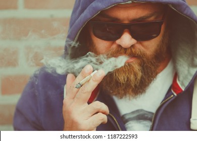 Young man in a hoodie with beard and sunglasses smoking marijuana joint - legalization (or legalisation) medical cannabis concept - weed or hashish cigarette with visible smoke from mouth