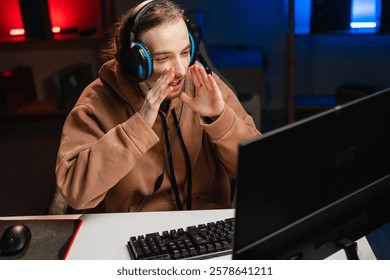 Young man in hoodie wearing headphones, sitting at desk, communicating excitedly while playing an online game, focused on monitor, illuminated by colorful gaming setup.