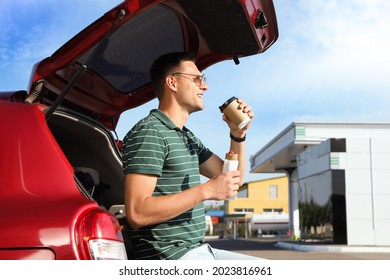 Young Man With Hot Dog Drinking Coffee Near Car At Gas Station