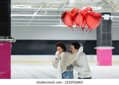 Young man hugging cheerful african american girlfriend with balloons in heart shape on ice rink  - Shutterstock ID 2232926805