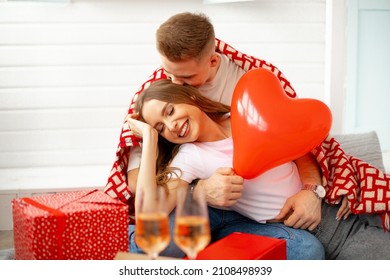 Young man hugs and kisses his beloved . They celebrate Valentine's Day at home in romantic setting with airy red hearts, gifts and wine.Love and care. Wonderful moments. Togetherness.