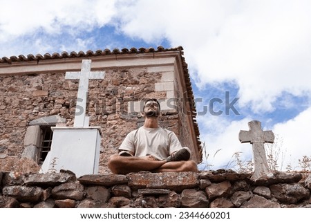 The young man of Indian origin practices Yoga next to two crosses in a stone church cemetery.