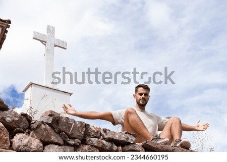 The young man of Indian origin stretches out his arms next to two crosses in a stone church cemetery. Christian faith