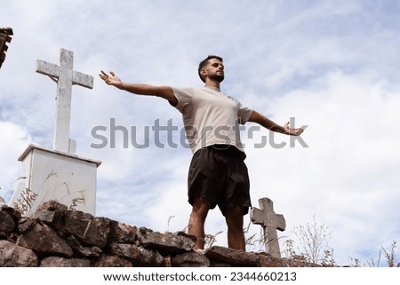 The young man of Indian origin takes a breath and stretches out his arms next to a cross in a stone church cemetery.