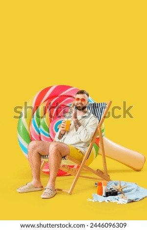 Young man with inflatable mattress in shape of lollipop and plastic cup of juice in deckchair on yellow background