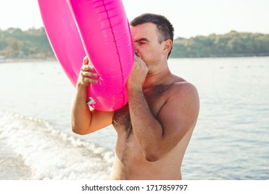 Young man inflates big pink inflatable ring on the summer beach. Summer vacation concept.