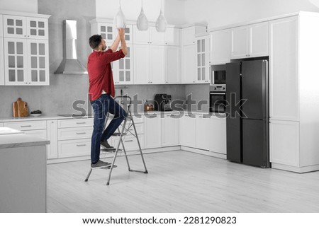 Young man installing ceiling lamp on stepladder in kitchen, space for text