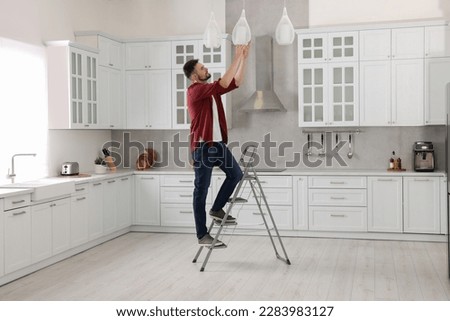 Young man installing ceiling lamp on stepladder in kitchen