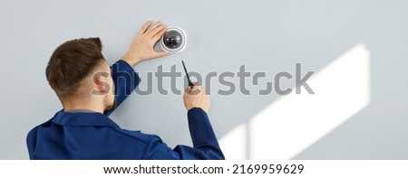 Young man installing modern security camera for theft deterrence inside new residential building. Repair service guy uses screwdriver to fit screws and adjust wall mounted CCTV surveillance dome cam
