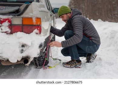 Young man is installing snow chains on a vintage car on snow and icy roads. Man struggling to get the car snow chains onto winter tires.
