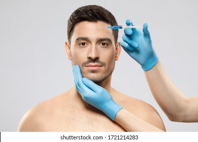 Young Man Isolated On Gray Background Looking At Camera While Receiving Botox Into Forehead To Prevent Sking Aging By Doctor In Blue Medical Gloves. Face Lifting Male