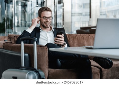 Young man it-worker CEO entrepreneur investor with suitcase sitting in modern airport waiting area while listening music using airpods, wireless headphone, working remote on laptop - Powered by Shutterstock