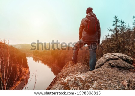 Young man in jacket, pants and backpack standing on rock looking to river Vltava and valley in sunset. 