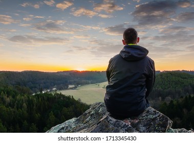 Young man in jacket, pants and  sitting on rock looking to valley at sunrise.