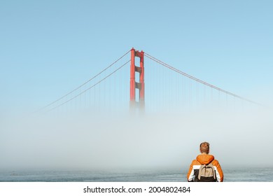 A young man with a jacket stands on the embankment and looks at the fog covering the Golden Gate Bridge in San Francisco, an incredible landscape