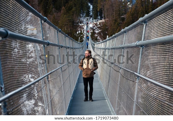 A young man in jacket stopped at the bridge\
during the hike, looking at the landscapes. Winter travel in\
Austria, Highline179.