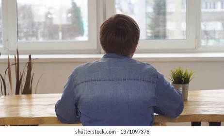 Young man in jeans shirt sitting with his back to the camera at the table near flower pot and doing something. Media. View from the back - Shutterstock ID 1371097376