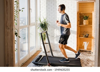 Young Man Jogging On The Modern Compact Treadmill At His Home At Room With Big Windows. Modern Lifestyle, Sport Indoors.