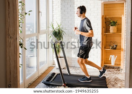 Young man jogging on the modern compact treadmill at his home at room with big windows. Modern lifestyle, sport indoors.