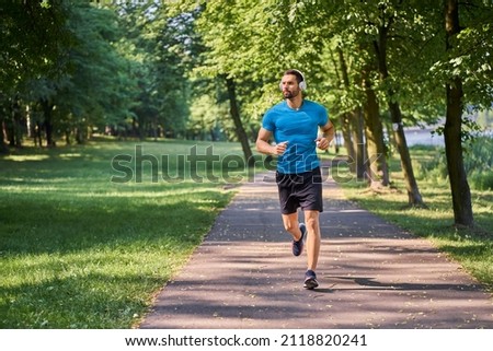 Young man jogging in a park on a sunny summer day listens to music on headphones