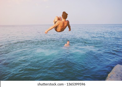 Young Man Jumping Off Cliff Into Blue Water Ocean At Sunset. Active Outdoor, Holiday Adventure, Tourism Action, Healthy Summer Joy, Fun Activity Lifestyle. Crazy Adult Guy In Swimwear Fly From Climb