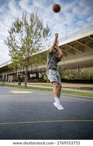 Young man jumping while shooting a basketball outdoors, practicing sports in a city park under a bridge