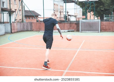 Young man kicks a football on an outdoor court, balancing his posture for a powerful shot. Wearing sporty attire, he plays in a vibrant setting with a goal and basketball hoop behind him. - Powered by Shutterstock