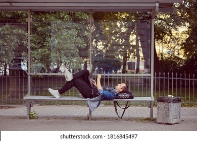 Young man with laptop on his knees is lying on the public transport station while waiting for a bus.