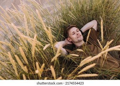 Young Man Laying In Beach Sand Grass. People Fatigue From Sunburn And Work. Summer Sleeping And Relaxation Techniques. Vitamin D Sunbathing. Man Power Nap With Eye Closed. Rest After Work From Home