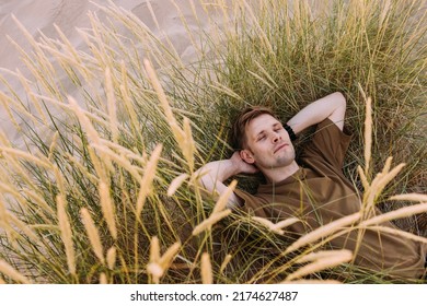 Young Man Laying In Beach Sand Grass. People Fatigue From Sunburn And Work. Summer Sleeping And Relaxation Techniques. Vitamin D Sunbathing. Man Power Nap With Eye Closed. Rest After Work From Home