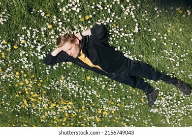 Young Man Laying In Green Grass With Flowers. People Fatigue From Work. Summer Sleeping And Relaxation Techniques. Vitamin D Sunbathing. Man Power Nap With Eye Closed. Rest After Work From Home