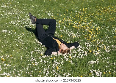 Young Man Laying In Green Grass With Flowers. People Fatigue From Work. Summer Sleeping And Relaxation Techniques. Vitamin D Sunbathing. Man Power Nap With Eye Closed. Rest After Work From Home