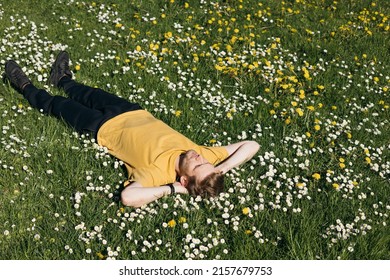 Young Man Laying In Green Grass With Flowers. People Fatigue From Work. Summer Sleeping And Relaxation Techniques. Vitamin D Sunbathing. Man Power Nap With Eye Closed. Rest After Work From Home