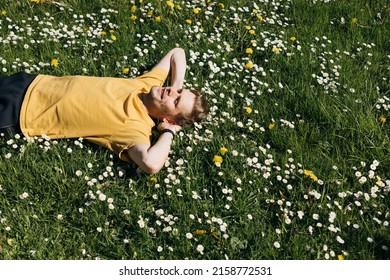 Young Man Laying In Green Grass With Flowers. People Fatigue From Work. Summer Sleeping And Relaxation Techniques. Vitamin D Sunbathing. Man Power Nap With Eye Closed. Rest After Work From Home