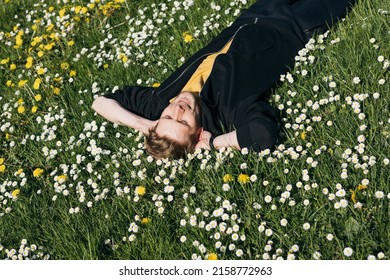 Young Man Laying In Green Grass With Flowers. People Fatigue From Work. Summer Sleeping And Relaxation Techniques. Vitamin D Sunbathing. Man Power Nap With Eye Closed. Rest After Work From Home