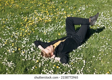 Young Man Laying In Green Grass With Flowers. People Fatigue From Work. Summer Sleeping And Relaxation Techniques. Vitamin D Sunbathing. Man Power Nap With Eye Closed. Rest After Work From Home