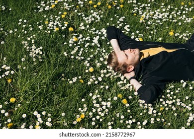 Young Man Laying In Green Grass With Flowers. People Fatigue From Work. Summer Sleeping And Relaxation Techniques. Vitamin D Sunbathing. Man Power Nap With Eye Closed. Rest After Work From Home