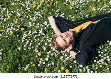 Young Man Laying In Green Grass With Flowers. People Fatigue From Work. Summer Sleeping And Relaxation Techniques. Vitamin D Sunbathing. Man Power Nap With Eye Closed. Rest After Work From Home