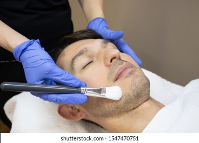 Young man laying on a facial therapy couch covered with the white towel with closed eyes. Facial therapist hands holding the facial black and white brush on a man face in a blue sterile gloves