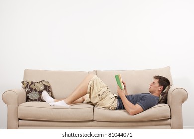 A Young Man Lays Down And Reads On A Couch In His Home.