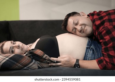 A young man leaned his ear against the pregnant belly of his wife, who was lying on her back on the sofa. Happy pregnant couple. A family is expecting a child. Conscious pregnancy concept - Shutterstock ID 2394559081