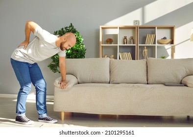 Young Man Leaning On Sofa At Home Is Holding His Lower Back Due To Severe Pain And Cramps. Concept Of Back Pain, Muscle Damage, Physical Injury, Sciatica, Arthritis, Rheumatism Or Nerve Entrapment.