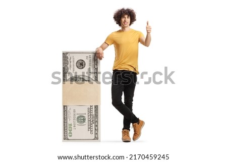 Young man leaning on a stack of money and showing thumbs up isolated on white background