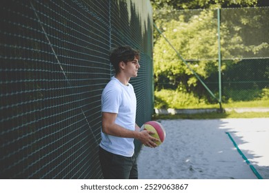 Young man leans against a fence holding a volleyball, looking thoughtfully into the distance. The scene captures a calm, reflective moment during a break on a sandy court, surrounded by greenery. - Powered by Shutterstock