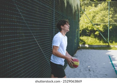 Young man leans against a fence holding a volleyball, looking thoughtfully into the distance. The scene captures a calm, reflective moment during a break on a sandy court, surrounded by greenery. - Powered by Shutterstock