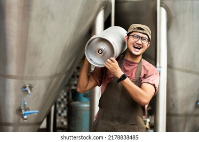 Young man in leather apron holding beer keg at modern brewery, craft brewery worker